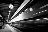 A train, blurred by the length of exposure, speeds past a platform, where a clock reads 6:48