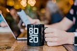 A person holds a mug placed on a desk while looking at their phone with their laptop in front of them.