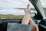 A young woman with her feet up on a car dashboard while looking at a map. The seashore is visible through the windshield. Photo by Leio McLauren on unsplash.com