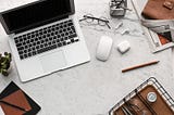 A laptop, computer mouse, a pair of air pods, a pair of reading glasses, and a pencil all sprawled out across a marble desk.