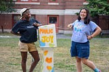 Sabrina and Ava stand with a four-foot-tall box between them on a patch of grass. They are both smiling, and Ava opens the top of the box. The box is painted with bright orange, yellow, green, and blue colors and says the words “Public Joy”.