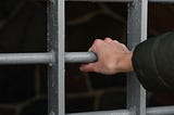 Metal jail cell door with white person’s hand gripping a bar. Unclear if the person is inside or outside of the cell.