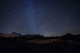 Mountains after dark, silhouetted against a dazzling night sky, with a smattering of glowing stars.