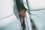 A young girl hiding behind a glass door with windswept hair.