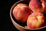 Three warm peaches lay in a cermaic bowl agaist a black background