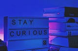A light box with the words “STAY CURIOUS” spelled out on it, sitting on a desk in front of a stack of books, with a pen lying on the desk in front of it. The entire photo has been tinted dark blue
