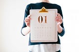 hands holding a clipboard with a month calendar on it and a big bold 01 in gold at the top of the paper on the clipboard