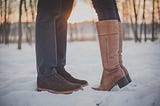 The legs and feet of a man and woman who appear to be kissing. The woman is standing on her tip toes in knee-high boots.