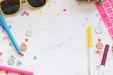 A Donut, markers, yellow sunglasses, coins and 6 dollars are sprawled uniformly on a white surface. the prominent color pink.