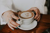 hands about to grasp a white mug of coffee with latte art on top of it