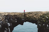 A person in a red rain jacket against a grey sky stands on jagged rocks spanning across water.