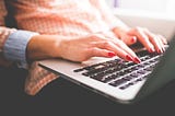 A white woman with red fingernail polish types on a Mac laptop, which sits on her lap.