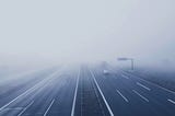 A multi-lane motorway disappearing into dense fog, with only a few vehicles visible and road markings fading into low visibility.