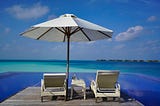 Two beach loungers on a desk, under a big white umbrella overlooking pristine blue waters.