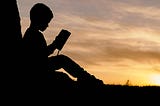 A silhouette of a child reading a book under a tree, against a yellow setting sun