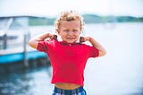 Image of toddler boy with curly sandy blonde hair wearing red short sleeve shirt and blue plaid shorts, with both arms flexed showing his strong muscles, standing on a dock with boats and the ocean in the background. The image was chosen by Spiritual Lifestyle Writer The Christine McDonald to portray a person’s superpower.