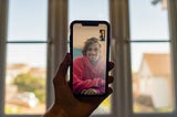 A hand holds a phone and the phone displays someone who is video chatting with the person on the other end of the hand — a blurry window and suburban scene with a house and car is in the background