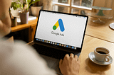 A person uses a laptop displaying the Google Ads logo on screen, seated at a wooden table with a cup of coffee nearby in a bright, cozy setting.