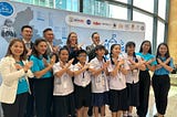 A group of adults and kids stand in front of a sign, posing with their wrists crossed in front of their chests.