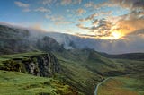 wide open vista, with craggy hills covered with green, a river in the distance, and sun peeking out behind clouds