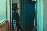 A woman in a black shirt and jeans with a striped undershirt leans against the doorframe of a rundown apartment.