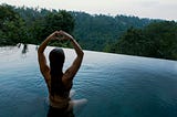 A woman in a bathing suit sitting in an infinity pool that is overlooking a lush green jungle. She is also making a heart sign with her two hands.