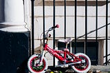 A pink child’s bike without training wheels propped against a wrought iron fence in a city