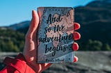 A hand is holding up a passport holder with the phrase “And so the adventure begins.” In the background is a blurred mountain terrain with a clear blue sky.