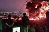 People silhoutted against the night sky, watching a huge fire against the dim lights of the city
