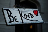 a hand-painted sign reads Be Kind with a red heart next to the word kind. black letters on a while background. wooden. hanging from something,maybe a door.