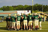 A team of high school football players in green and gold uniforms huddle up around the 50 yard line.