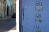 An exterior building wall painted blue has the phrase “GRL PWR” written vertically three times. To the left of the blue wall is an out-of-focus street or alleyway in an urban setting.