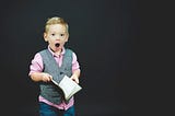 Young boy holding a book looks shocked at something he has read.