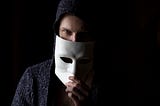 a man in the dark holds up a white mask to cover half his face