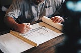An architect at work sits at a wooden desk drawing a sketch.