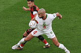 Two men playing soccer on a field, kicking the ball towards each other with determination.