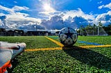 A soccer ball with a star is placed on yellow lines on a soccer field. A cleat at the bottom left of the photograph prepares to kick the ball toward the goal in the distance. Puffy cumulous clouds rise over the field with a bright shining sun and brilliant blue sky.