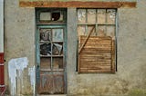 A photo of the front of a house, with a broken door and a broken window