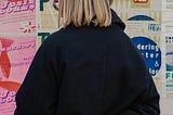 A woman with short blond hair wearing a black trench coat, crossing her fingers behind her back. She is standing in front of a colourful wall.