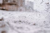 A snow-spackled window shows a blurry display of snow carpeting the ground and a large brown building in the far background.