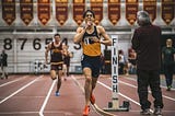 A runner finishes a race at on indoor track meet and appears to be silencing the crowd with his index finger over his lips.
