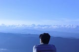 A young man sits on a hilltop, facing away and looking out over a series of hills and valleys.