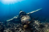 Underwater war plane, on the ocean floor. It’s partially eroded, with algae growing on top. Light is shining from top left