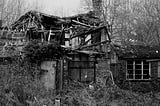 A black and white picture of an old, decaying home or barn. Rafters are exposed and toppling over. The building is barely still standing.