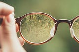 A hand holding a glasses lens focusing on the lush greenery view