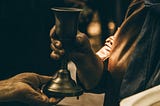 Cult member in a rope hands a glass of liquid to a follower