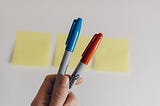 A blue and orange permanent marker is held up to camera in front of two post-it notes