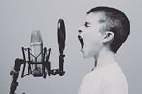 Black and white photo of a young boy with short hair screaming into a large microphone.