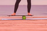 An image of legs from the knees down wearing black leggings and bare feet balancing on a board that is on a round green cylinder.