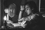 A thoughtful man sits in front of a bare bulb and a desk fan, rereading his writing in a notebook.